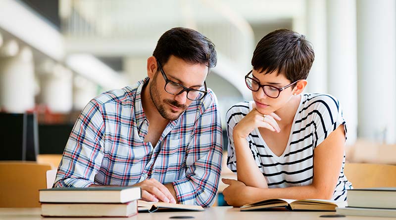 Um professor com óculos, camisa xadrez, lendo um livro para a aluna, que também esta de óculos, e uma blusa listrada, apoiada em um livro aberto