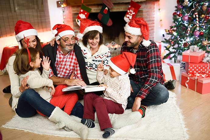Uma família, quatro adultos, com gorro de papai Noel, duas crianças, sentados em um tapete no chão, ao lado de uma arvore de natal, e presentes