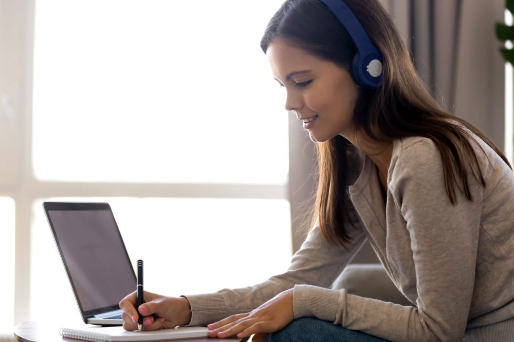 Uma mulher, com fone de ouvido, de frente para o notebook, com uma caneta na mão fazendo anotações